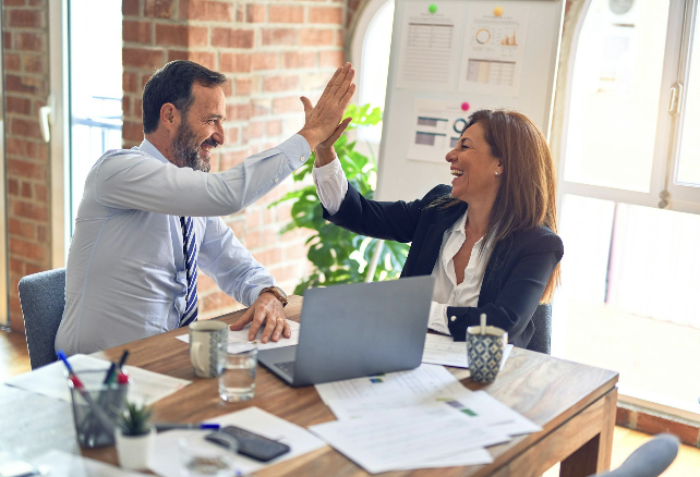 Two colleagues celebrating with a high five across a desk in a bright office.